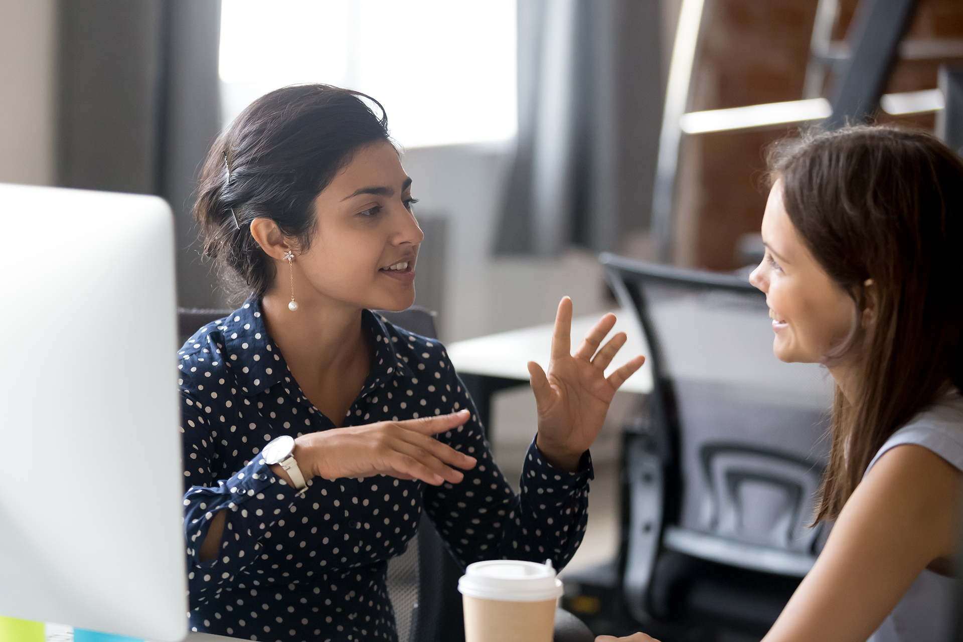 Friendly female colleagues having good relationships, pleasant conversation at workplace during coffee break, smiling young woman listen talkative coworker, discussing new project, talking in office