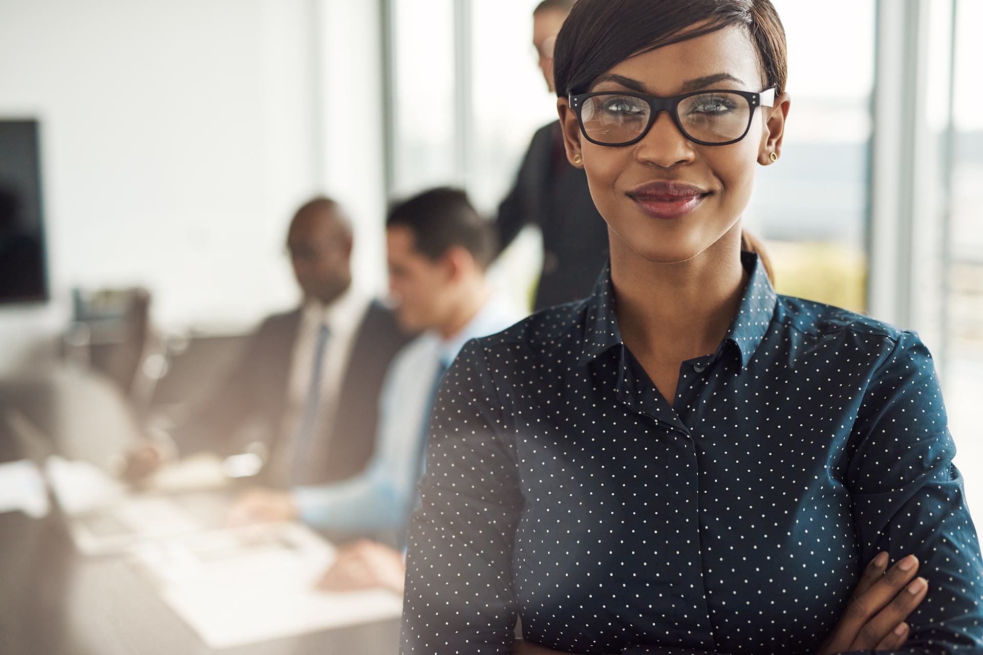 Beautiful young grinning professional Black woman in office with eyeglasses, folded arms and confident expression as other workers hold a meeting in background